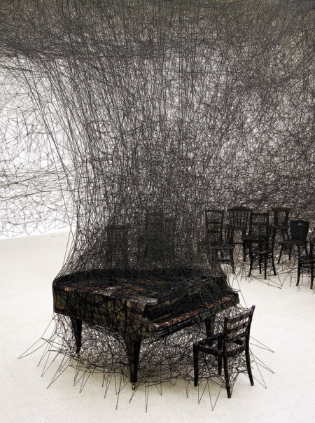 chiharushiotainsilence2 When I first set my eyes on Japan-born, Berlin-based artist Chiharu Shiota's work, I wasn't sure if I was looking at an installation or a dark charcoal illustration. Though the piece echoes sketch-like imagery, it is in fact an installation piece involving a burnt piano in a room ravaged by black wool. The work known as In Silence is inspired by Shiota's own traumatic memories as a child, having witnessed her neighbor's house burn down. The charred piano is a direct memory of her neighbor's grand piano blazed up in smoke.  There is a melancholic aura that hovers throughout the incinerated room filled with singed furniture. The miles of thread woven in, around, and through each item within the space adds a feeling of entrapment. The way it engulfs the room's furnishings encapsulates the destructive and overwhelming nature of flames that have possessed one's material properties.